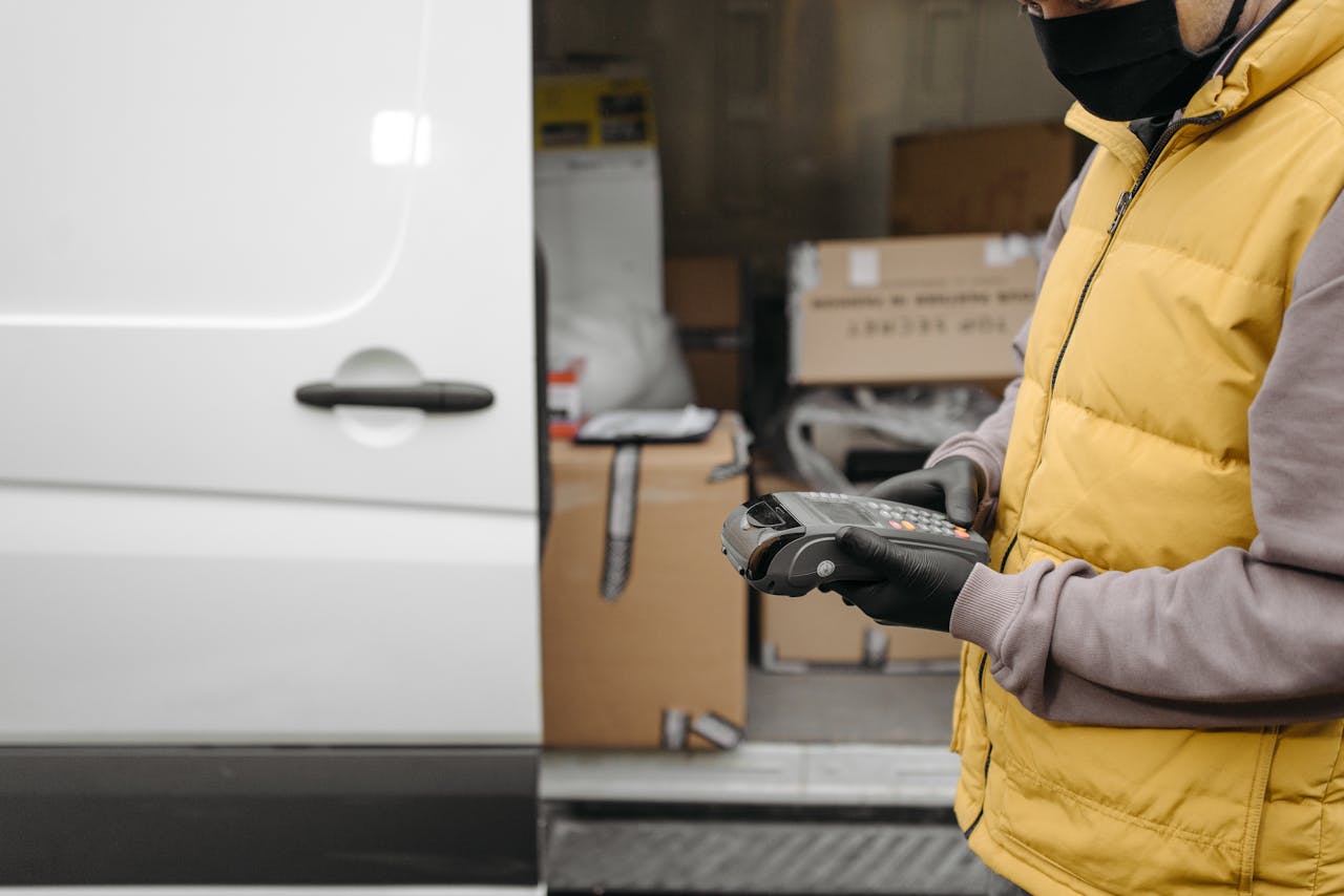 Courier using a terminal for contactless delivery payment next to a van with packages.